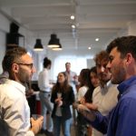 Two professionals in a focused one-on-one conversation at a business networking event, with a small group talking in the background
