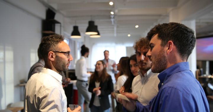 Two professionals in a focused one-on-one conversation at a business networking event, with a small group talking in the background