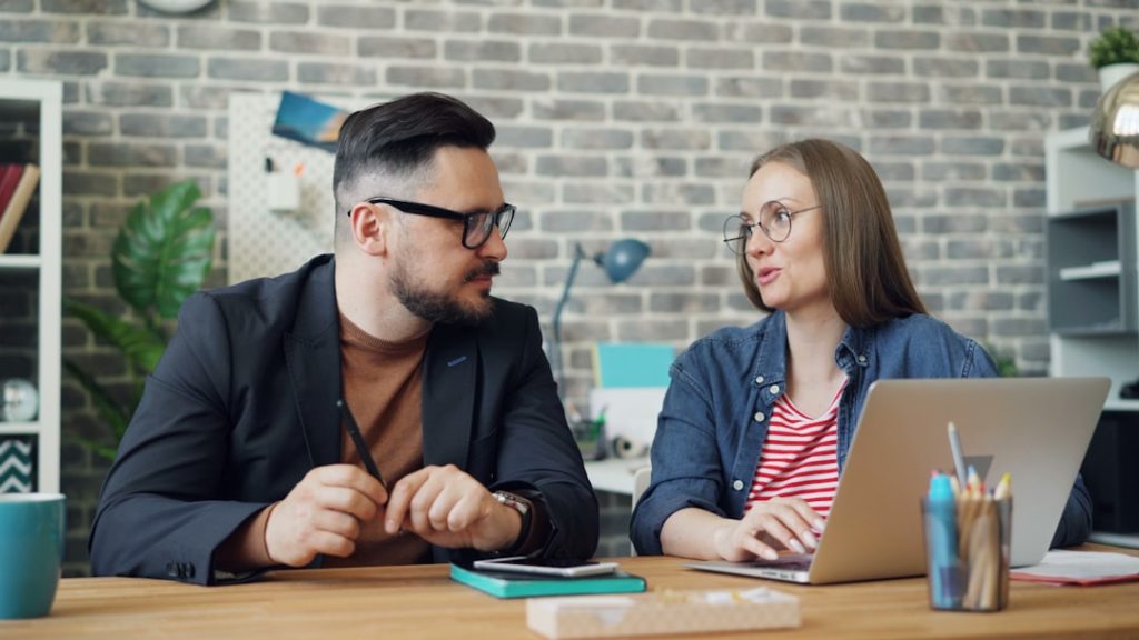 Two people in a one-on-one working session at a desk, talking over an open laptop and notebook, the kind of check-in a coach runs with a client.