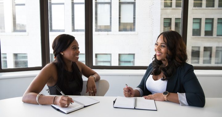 Two women in conversation across a table, each holding a pen, in the kind of one-to-one meeting a coaching practice is built on.