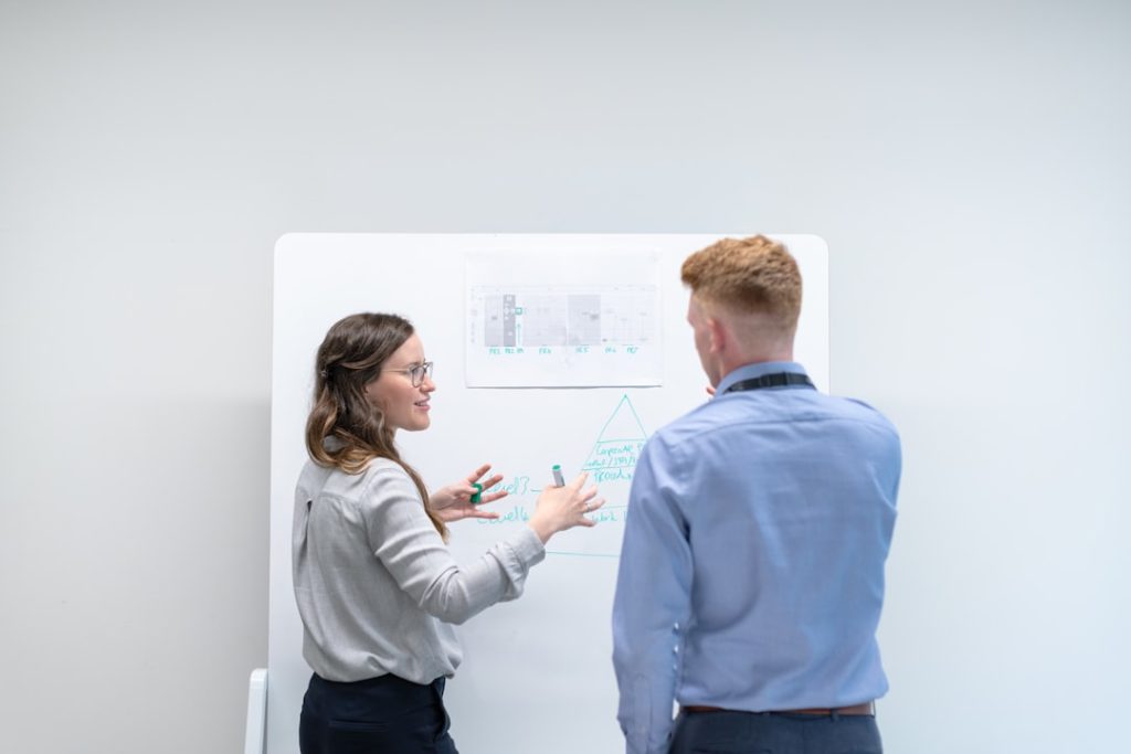 A woman in a grey shirt gestures toward a whiteboard as she explains a hand-drawn diagram to a colleague in a light blue shirt, the two of them standing one-on-one in a plain white meeting room.