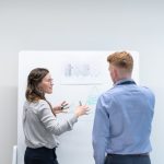 A woman in a grey shirt gestures toward a whiteboard as she explains a hand-drawn diagram to a colleague in a light blue shirt, the two of them standing one-on-one in a plain white meeting room.