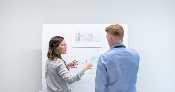 A woman in a grey shirt gestures toward a whiteboard as she explains a hand-drawn diagram to a colleague in a light blue shirt, the two of them standing one-on-one in a plain white meeting room.