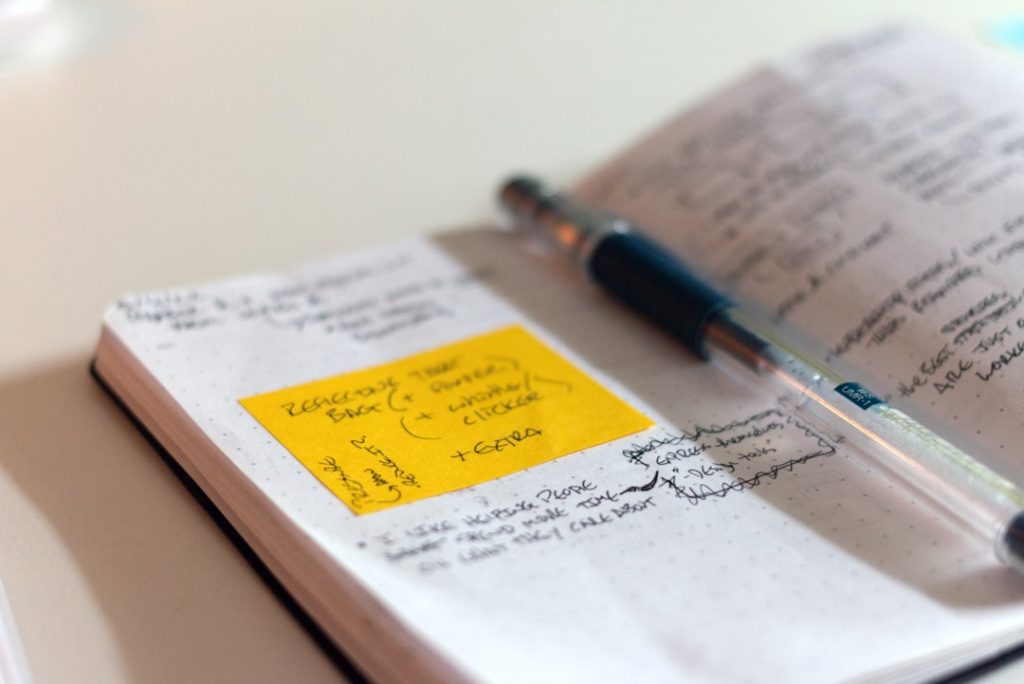 An open dot-grid notebook covered in handwritten coaching notes, with a yellow sticky note of reminders stuck to the left page and a ballpoint pen lying across the spine.