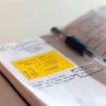 An open dot-grid notebook covered in handwritten coaching notes, with a yellow sticky note of reminders stuck to the left page and a ballpoint pen lying across the spine.