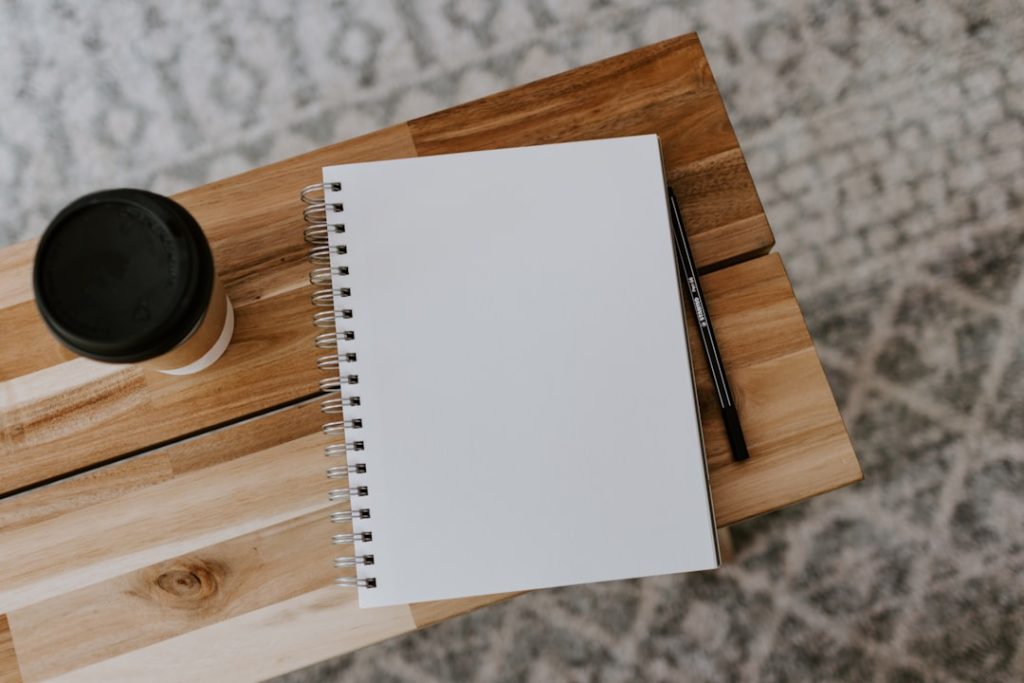An open blank spiral notebook and a black pencil on a light wooden table, photographed from above, with a lidded takeaway coffee cup to the left and a patterned grey rug beneath.