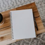 An open blank spiral notebook and a black pencil on a light wooden table, photographed from above, with a lidded takeaway coffee cup to the left and a patterned grey rug beneath.