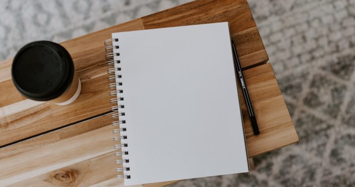 An open blank spiral notebook and a black pencil on a light wooden table, photographed from above, with a lidded takeaway coffee cup to the left and a patterned grey rug beneath.