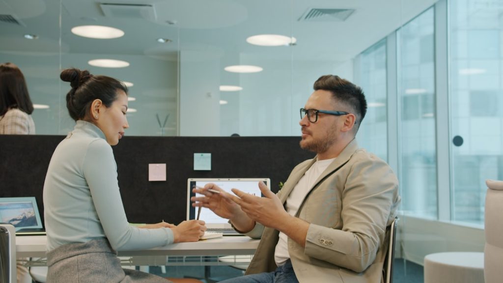 A woman in a light grey turtleneck takes handwritten notes beside an open laptop while a man in a beige blazer gestures mid-sentence across a white desk in a bright modern office.