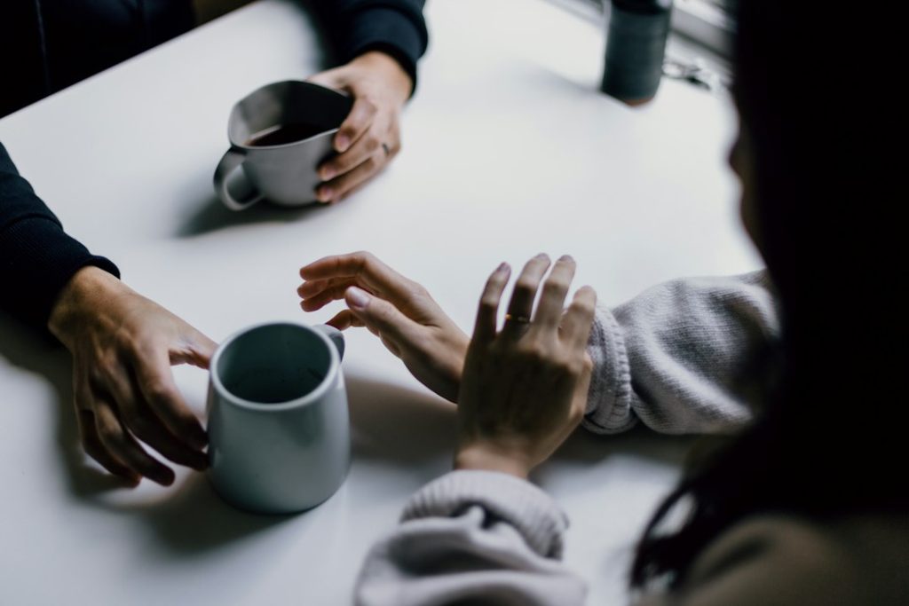Overhead view of two people talking across a white table, one holding a grey mug of coffee, the other in a cream sweater gesturing with both hands mid-conversation
