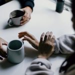 Overhead view of two people talking across a white table, one holding a grey mug of coffee, the other in a cream sweater gesturing with both hands mid-conversation
