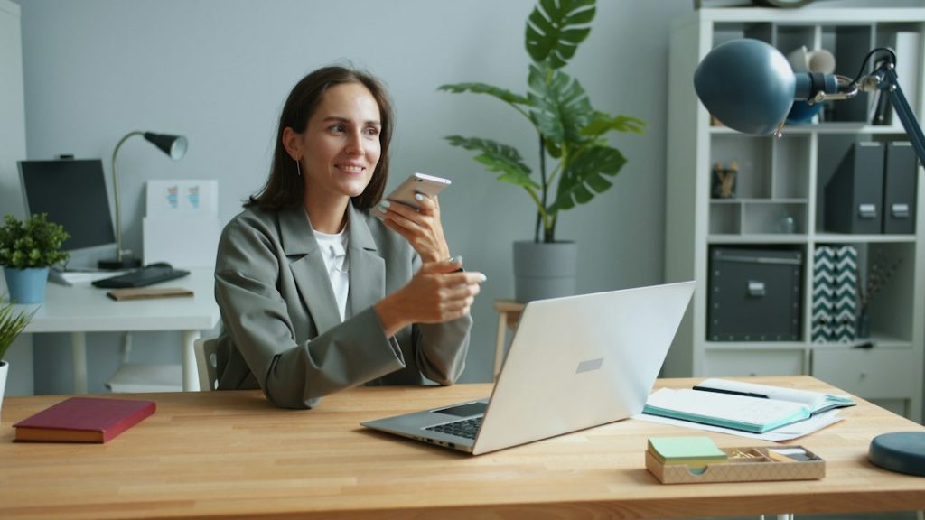 A smiling woman in a grey blazer sits at a wooden desk in a bright office, holding a smartphone up near her face as if recording a voice note, with an open laptop, a notebook, and a potted plant beside her.