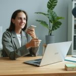 A smiling woman in a grey blazer sits at a wooden desk in a bright office, holding a smartphone up near her face as if recording a voice note, with an open laptop, a notebook, and a potted plant beside her.
