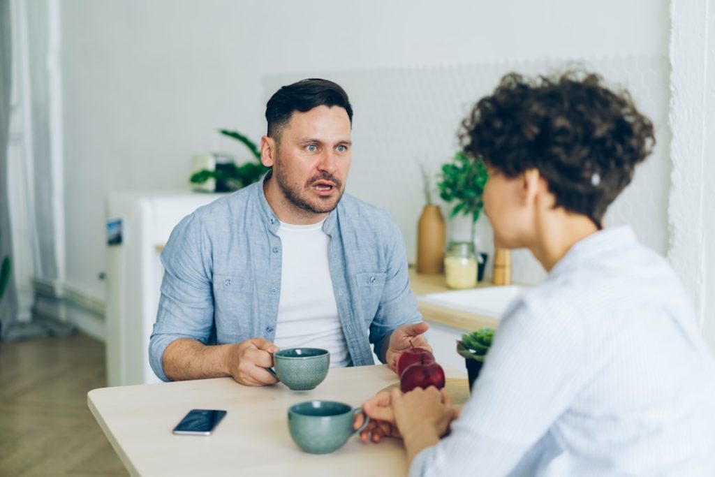 Two people sit across a pale wooden table in a bright room, each holding a mug, talking one on one mid-conversation.