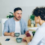 Two people sit across a pale wooden table in a bright room, each holding a mug, talking one on one mid-conversation.