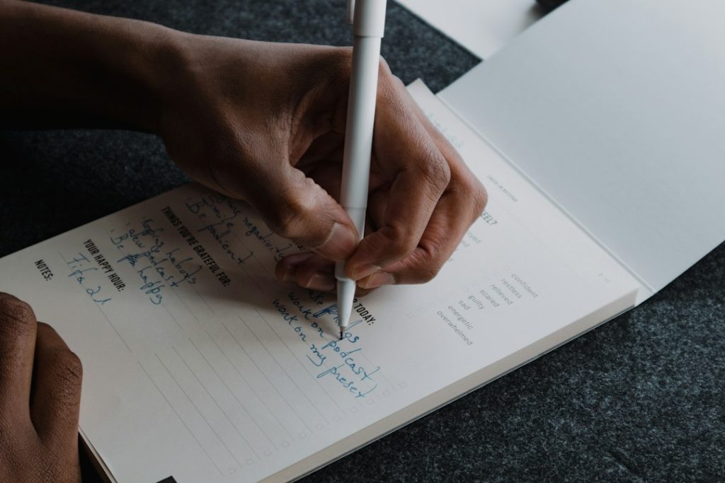 Close-up of a hand writing with a white pen in a printed daily check-in journal, filling in a to-do line beside a column of mood checkboxes
