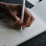 Close-up of a hand writing with a white pen in a printed daily check-in journal, filling in a to-do line beside a column of mood checkboxes