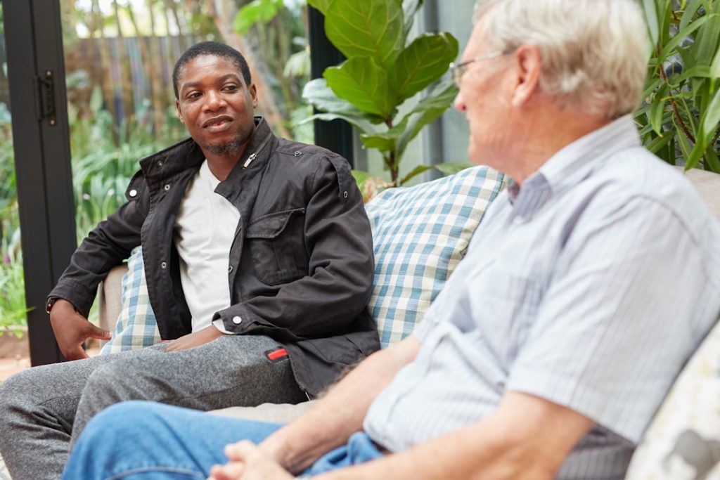 Two men in conversation on a couch with blue-and-white checked cushions in a bright sunroom with large green plants: a younger man in a dark jacket and white t-shirt speaks while an older man with white hair and glasses, in a striped shirt, listens.
