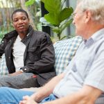 Two men in conversation on a couch with blue-and-white checked cushions in a bright sunroom with large green plants: a younger man in a dark jacket and white t-shirt speaks while an older man with white hair and glasses, in a striped shirt, listens.