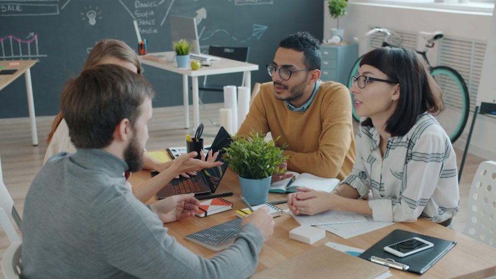 Four colleagues talking around a wooden table in a bright office, one with an open laptop and notebook, a chalkboard sketch of a plan behind them