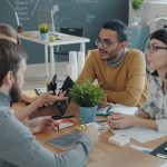 Four colleagues talking around a wooden table in a bright office, one with an open laptop and notebook, a chalkboard sketch of a plan behind them