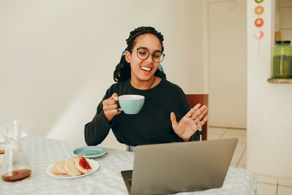 A smiling woman in glasses holds a teal coffee cup and waves at her laptop during a video call at a kitchen table.