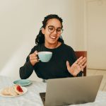 A smiling woman in glasses holds a teal coffee cup and waves at her laptop during a video call at a kitchen table.