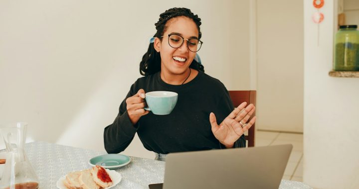 A smiling woman in glasses holds a teal coffee cup and waves at her laptop during a video call at a kitchen table.