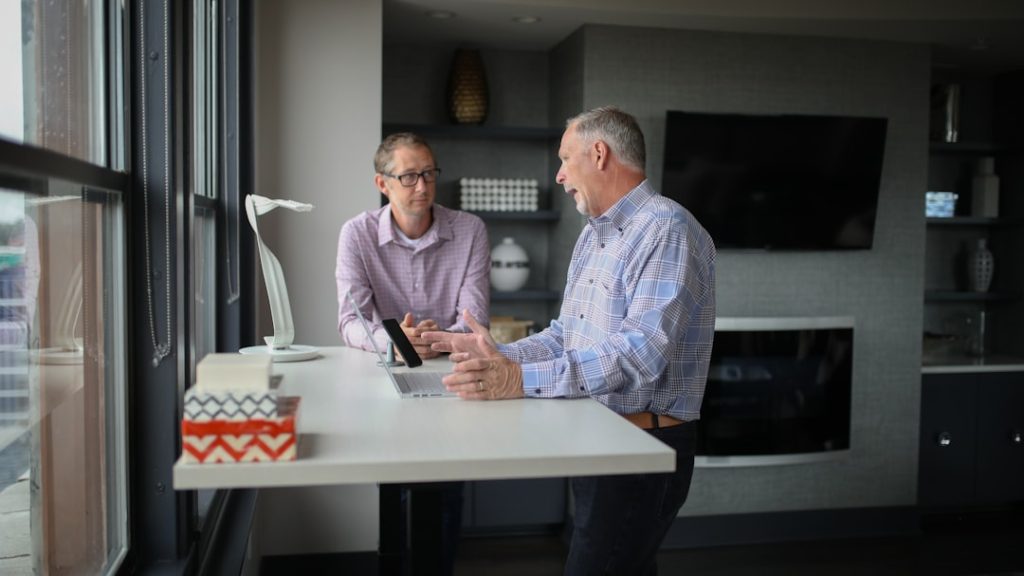Two men in plaid shirts talking across a white standing desk by a window, one gesturing mid-explanation over an open laptop, the other listening with hands folded, in a grey modern office.