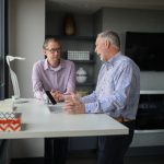 Two men in plaid shirts talking across a white standing desk by a window, one gesturing mid-explanation over an open laptop, the other listening with hands folded, in a grey modern office.