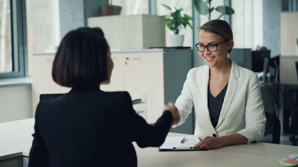 A smiling woman in glasses and a white blazer shakes hands across an office desk with another woman, a clipboard with a signed paper and pen in front of her