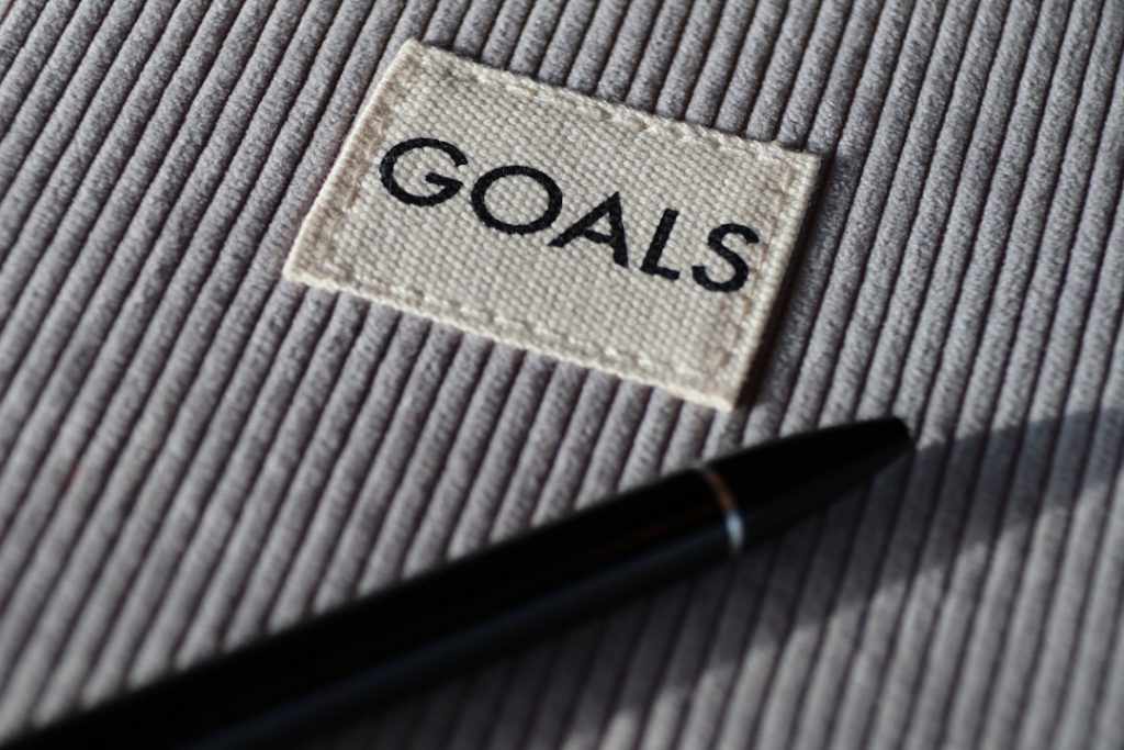 Close-up of a grey ribbed notebook cover with a woven label reading GOALS and a black pen resting across it
