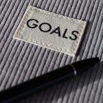 Close-up of a grey ribbed notebook cover with a woven label reading GOALS and a black pen resting across it