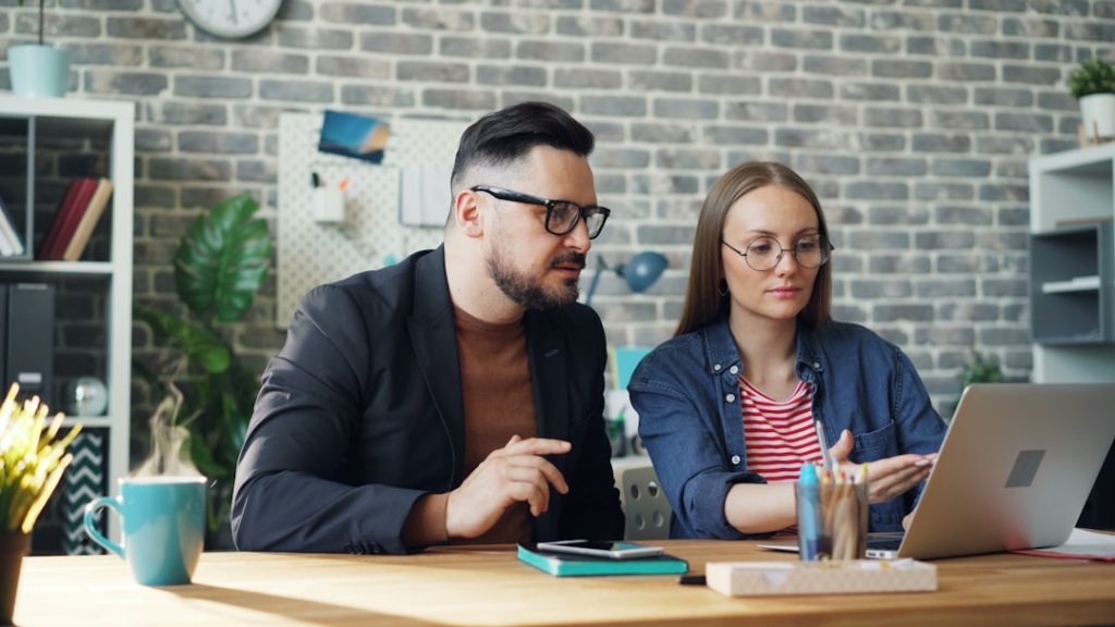 A man in glasses and a dark blazer and a woman in a denim shirt sit side by side at a wooden desk, reviewing something on an open laptop together, in a bright office with a grey brick wall, a teal mug, and a notebook and phone on the desk.