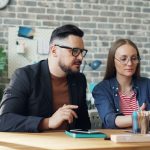 A man in glasses and a dark blazer and a woman in a denim shirt sit side by side at a wooden desk, reviewing something on an open laptop together, in a bright office with a grey brick wall, a teal mug, and a notebook and phone on the desk.