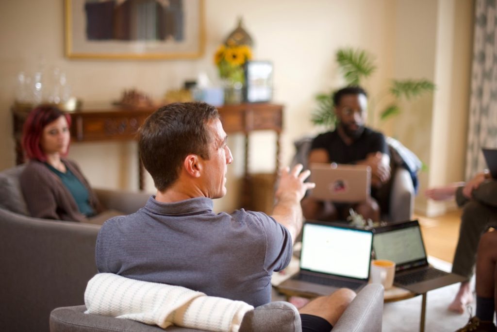 Four people sitting in armchairs around a low table in a sunlit living room, laptops open, one man gesturing mid-sentence while the others listen.