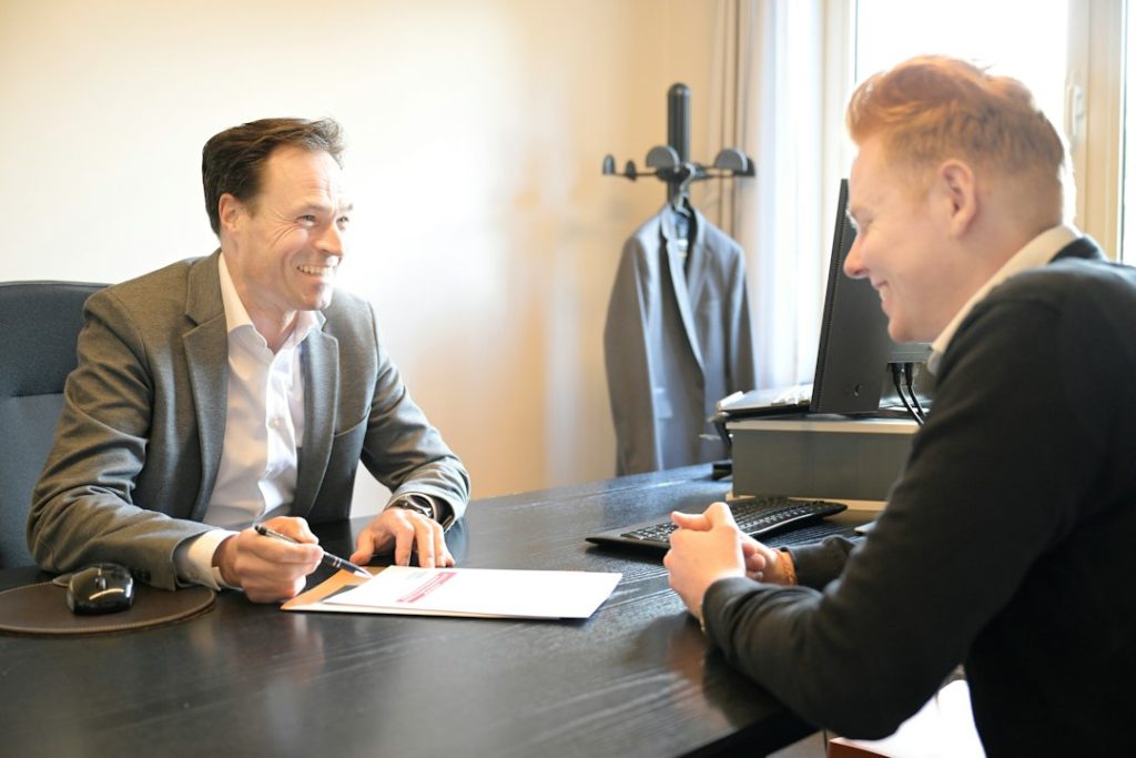 Two smiling men in a one-on-one meeting at a black office desk, one in a grey blazer holding a pen over a printed document while the other leans in to talk