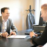 Two smiling men in a one-on-one meeting at a black office desk, one in a grey blazer holding a pen over a printed document while the other leans in to talk