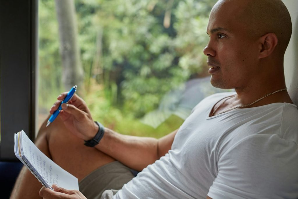 A man in a white t-shirt sits by a window with greenery outside, holding a pen and a handwritten notebook page while looking away in thought, reviewing his notes.