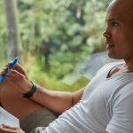 A man in a white t-shirt sits by a window with greenery outside, holding a pen and a handwritten notebook page while looking away in thought, reviewing his notes.