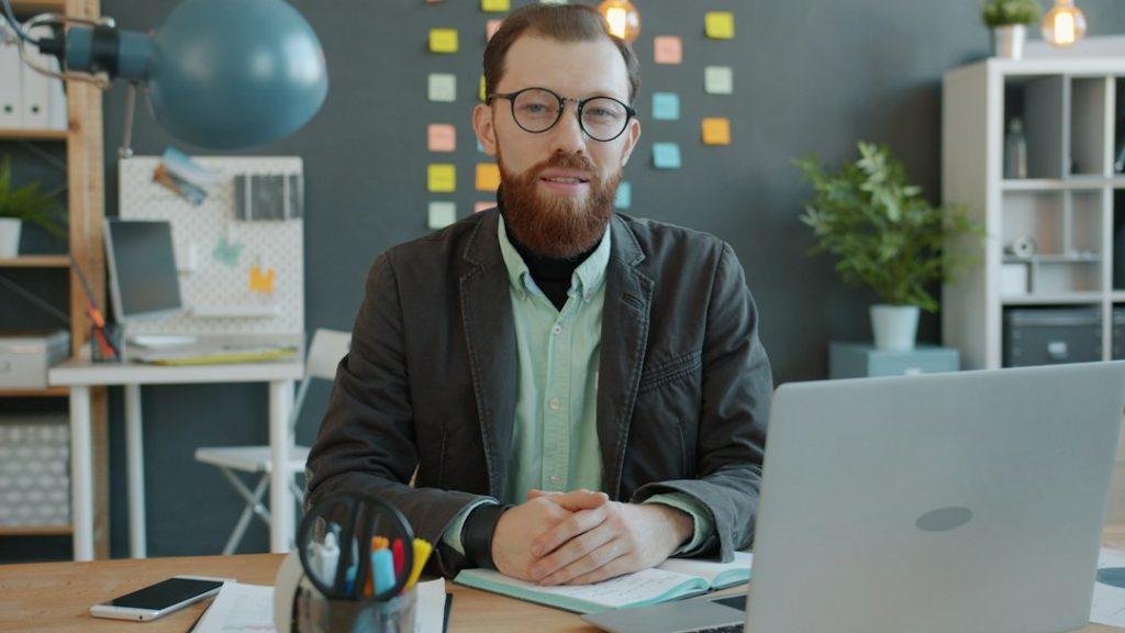 A bearded coach in glasses and a grey jacket sits at a wooden desk with an open notebook and a laptop, hands clasped, in front of a dark wall covered in colourful sticky notes