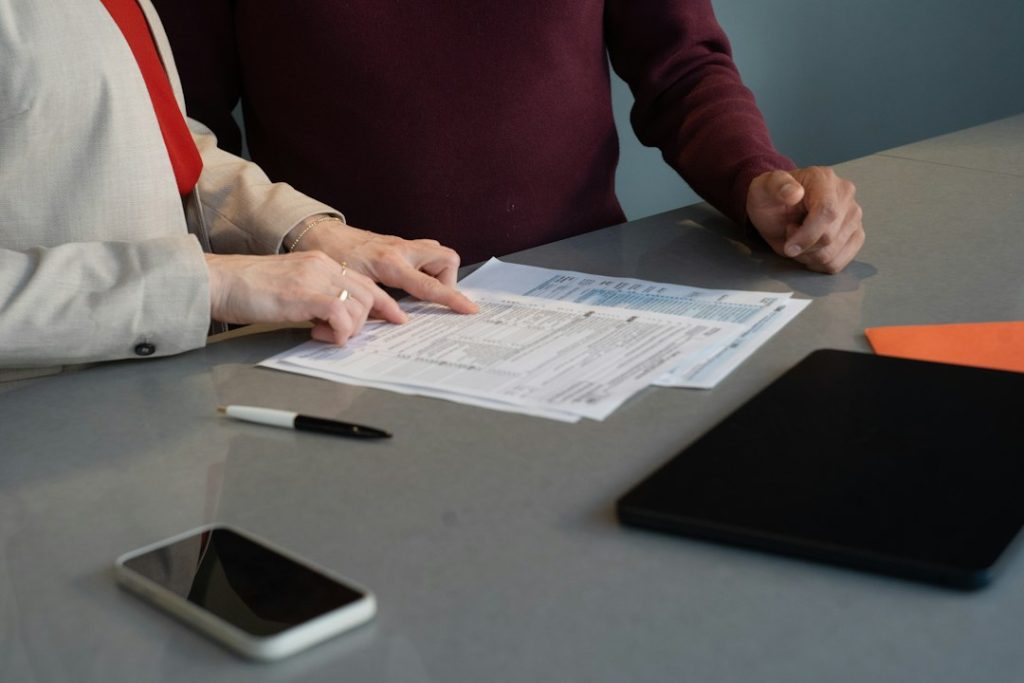 Two people at a grey table, one pointing to a line on a printed form, with a pen, phone and tablet beside the papers