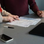 Two people at a grey table, one pointing to a line on a printed form, with a pen, phone and tablet beside the papers