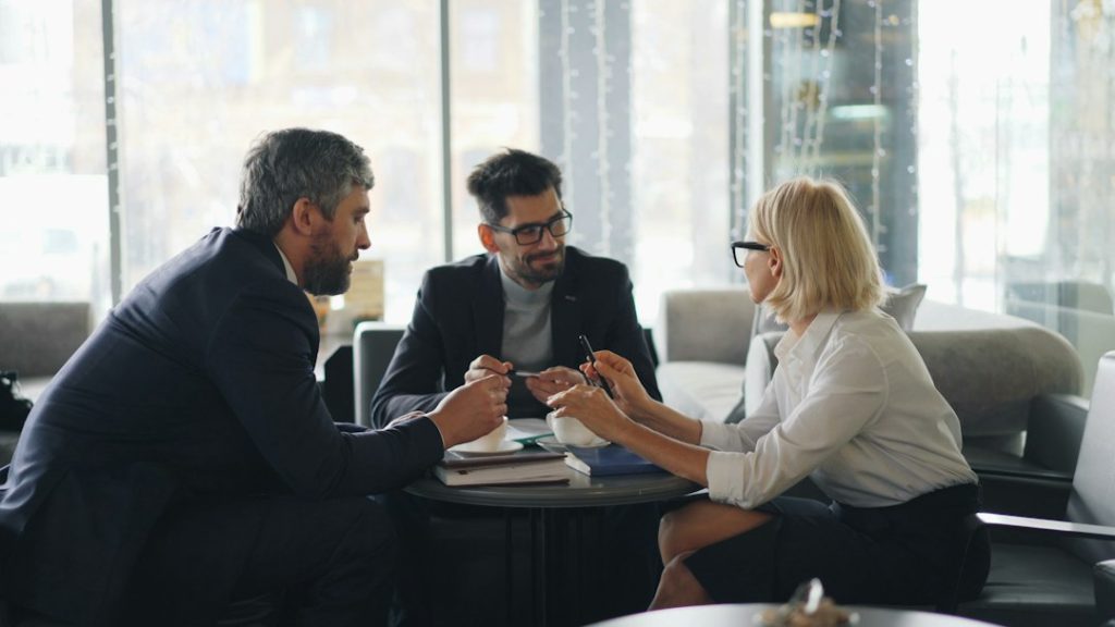 Three professionals talk over coffee at a small round table in a bright cafe, one woman in glasses holding a pen above a notebook