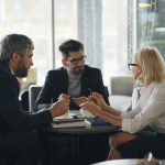 Three professionals talk over coffee at a small round table in a bright cafe, one woman in glasses holding a pen above a notebook