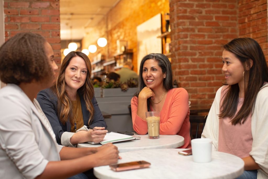 Four people seated around a small round marble cafe table in conversation, one holding a pen over an open spiral notebook while the others lean in listening, with an exposed brick wall and warm pendant lighting behind them.