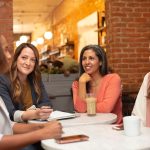 Four people seated around a small round marble cafe table in conversation, one holding a pen over an open spiral notebook while the others lean in listening, with an exposed brick wall and warm pendant lighting behind them.