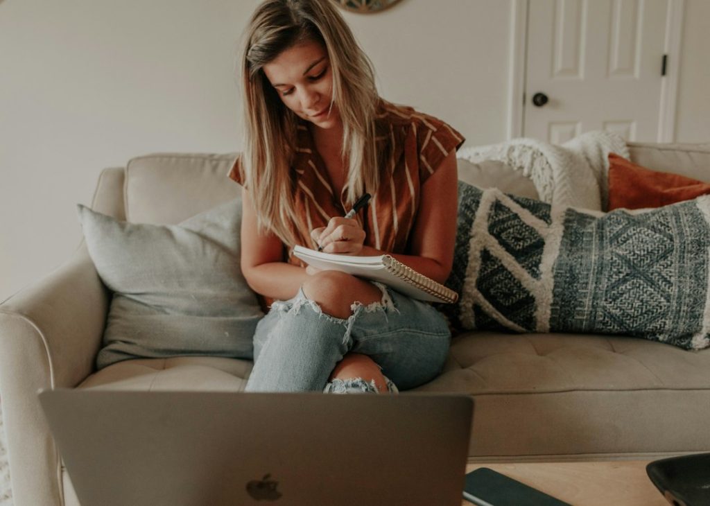 A woman in a rust striped shirt and ripped jeans sits cross-legged on a beige sofa, writing in a spiral notebook, with an open laptop in the foreground and patterned cushions beside her.