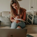 A woman in a rust striped shirt and ripped jeans sits cross-legged on a beige sofa, writing in a spiral notebook, with an open laptop in the foreground and patterned cushions beside her.
