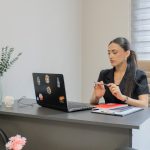 A coach sits at a grey desk in a bright office, pen in hand, reviewing an open laptop with a stack of notebooks and a red notepad beside her and an empty chair across the desk.